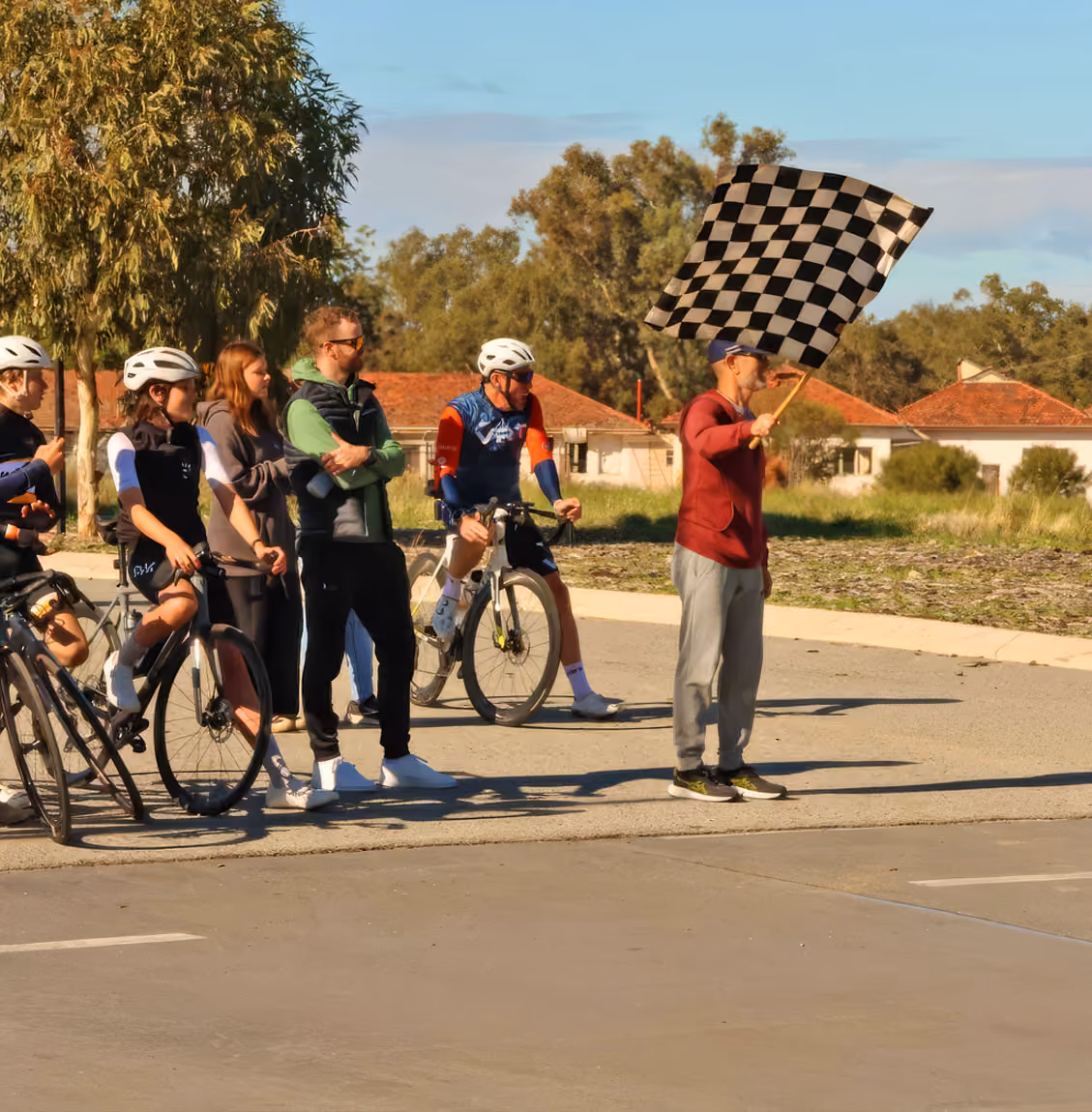 Person waving a black-and-white checkered flag as cyclists and spectators stand nearby on a sunny day.
