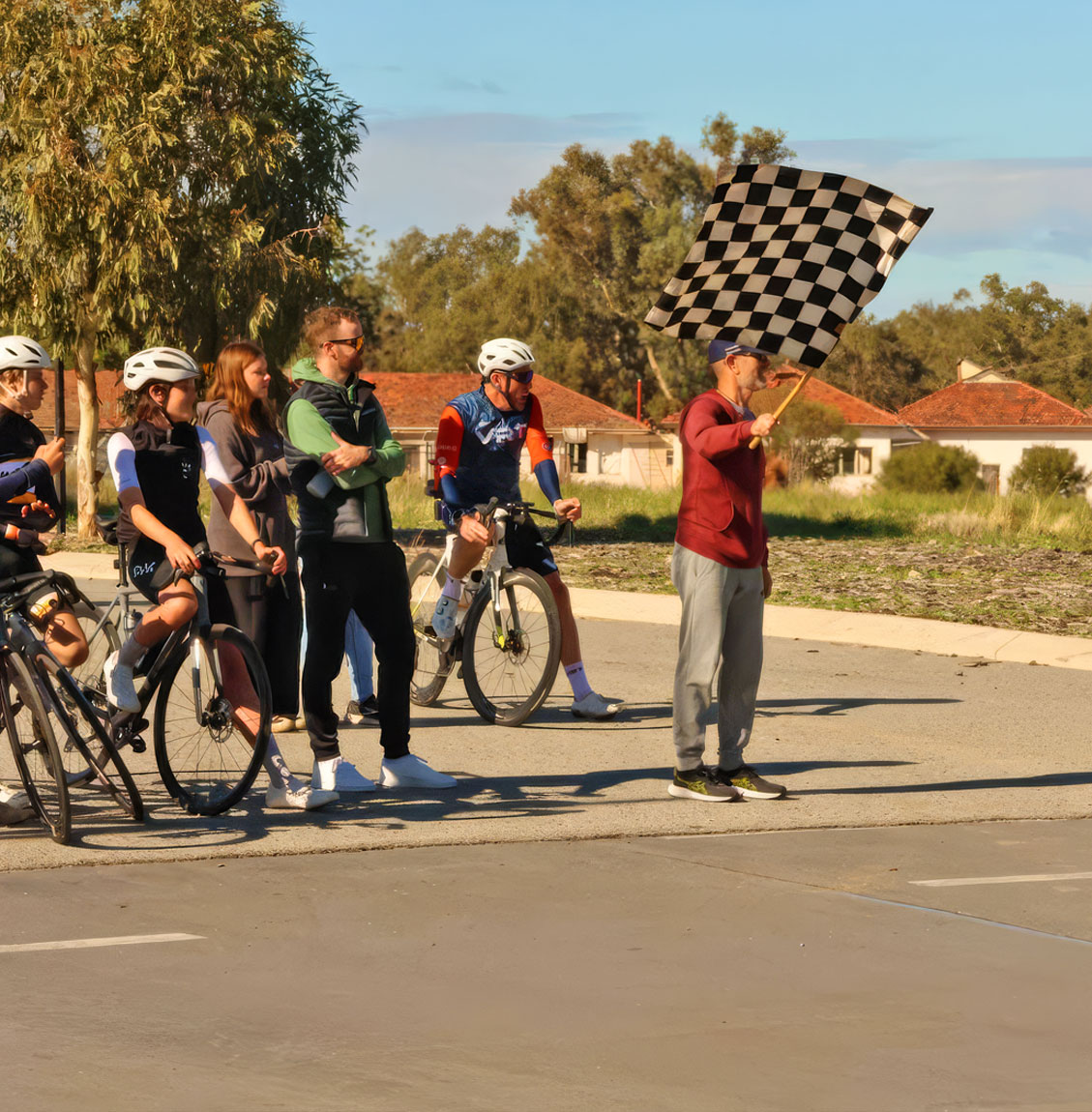 Person waving a black-and-white checkered flag as cyclists and spectators stand nearby on a sunny day.