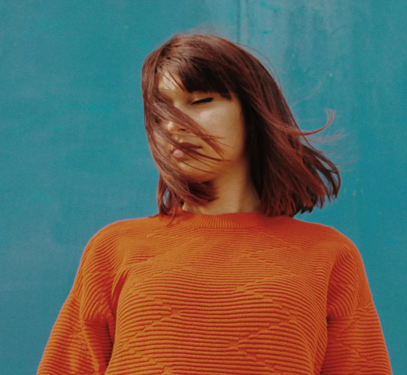 Woman with short brown hair wearing an orange sweater standing against a blue background, hair blowing across her face.