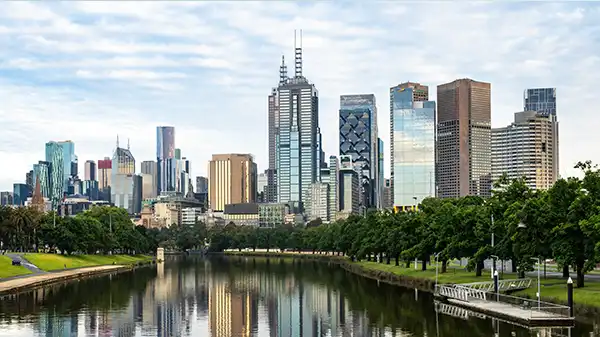 Melbourne skyline reflected in calm river with green trees and cloudy sky above.
