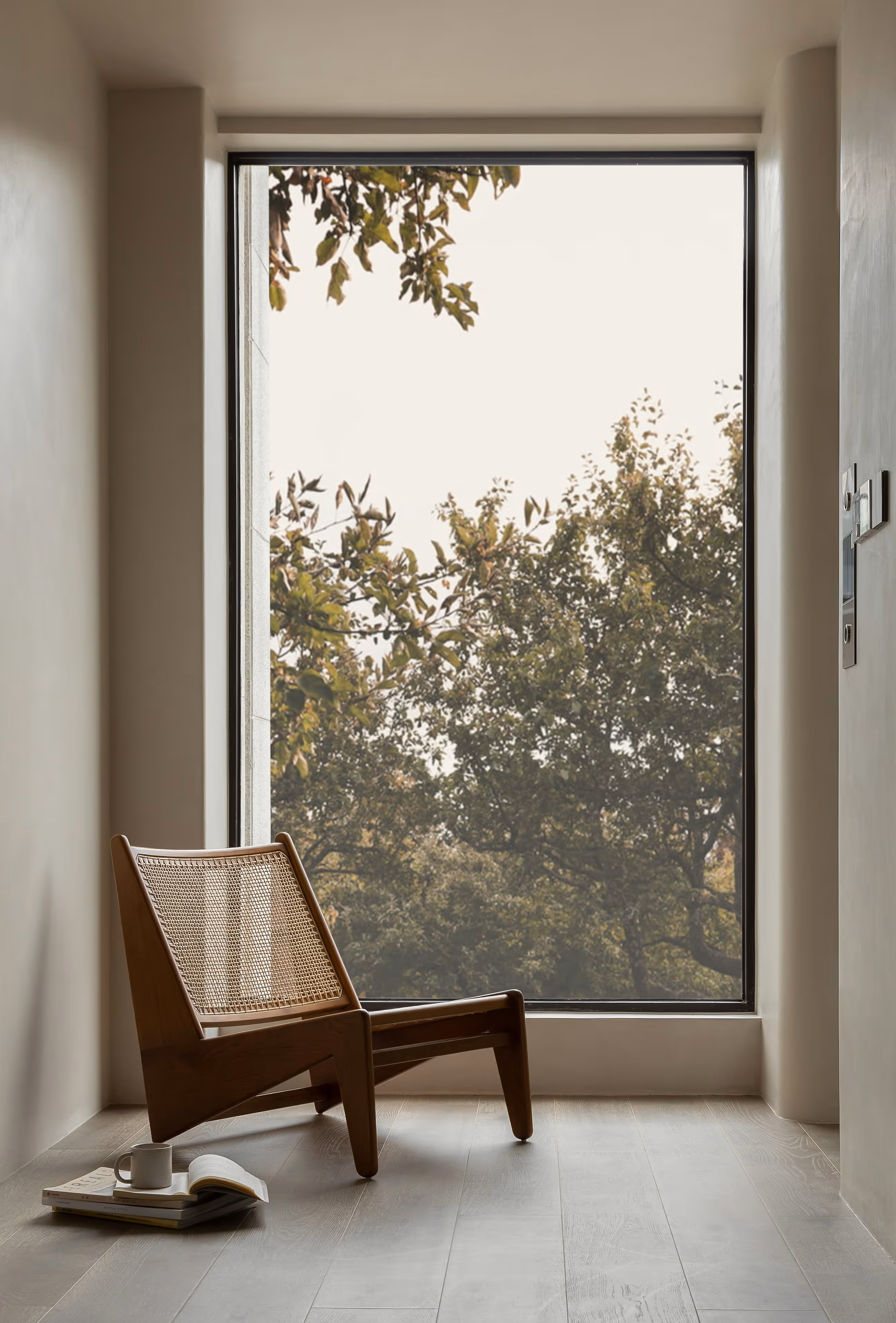 Wooden lounge chair with woven back by a large window showing tree foliage outside on wooden floor.