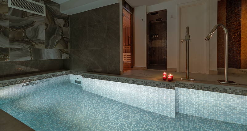 Indoor spa pool with mosaic tiles, soft lighting, candles, and adjacent sauna and shower rooms.