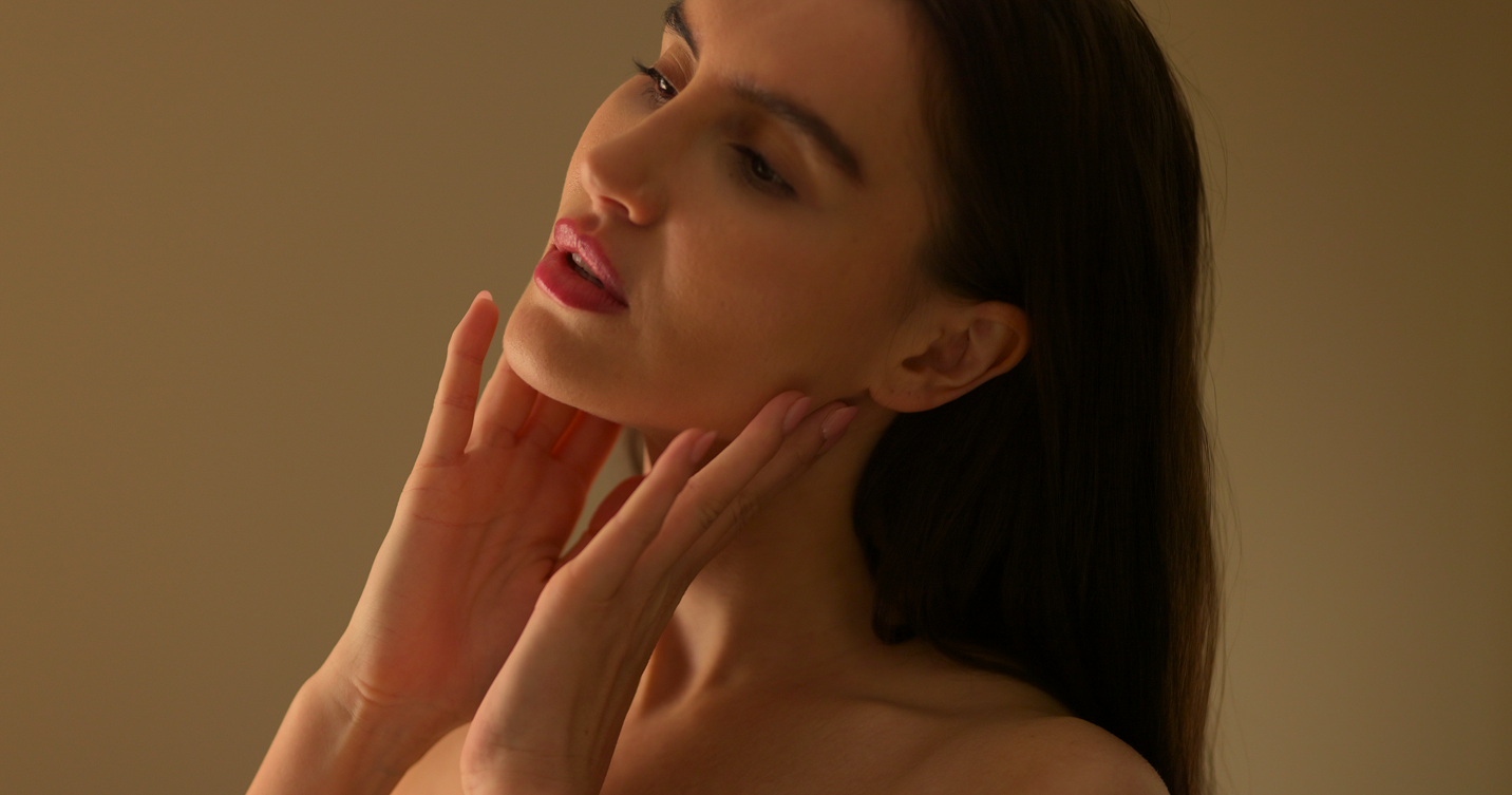 Woman with long dark hair touching her neck and chin against a plain beige background.