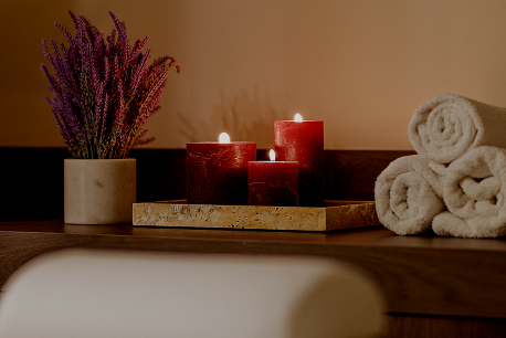 Three red lit candles on a tray beside lavender flowers and rolled white towels on a wooden surface.