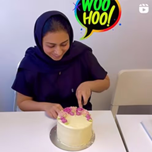 Woman in dark clothing and headscarf cutting a white cake decorated with pink roses on a white table.