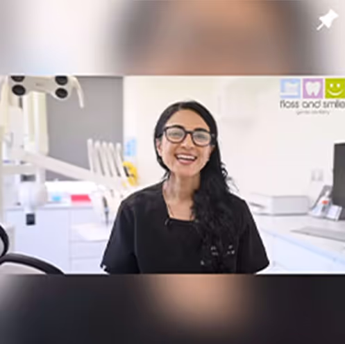Smiling woman with long dark hair and glasses in black scrubs sitting in a bright dental office.