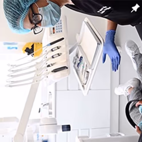 Dentist wearing a surgical mask, hair cover, glasses, and blue gloves examining a patient in a dental clinic.