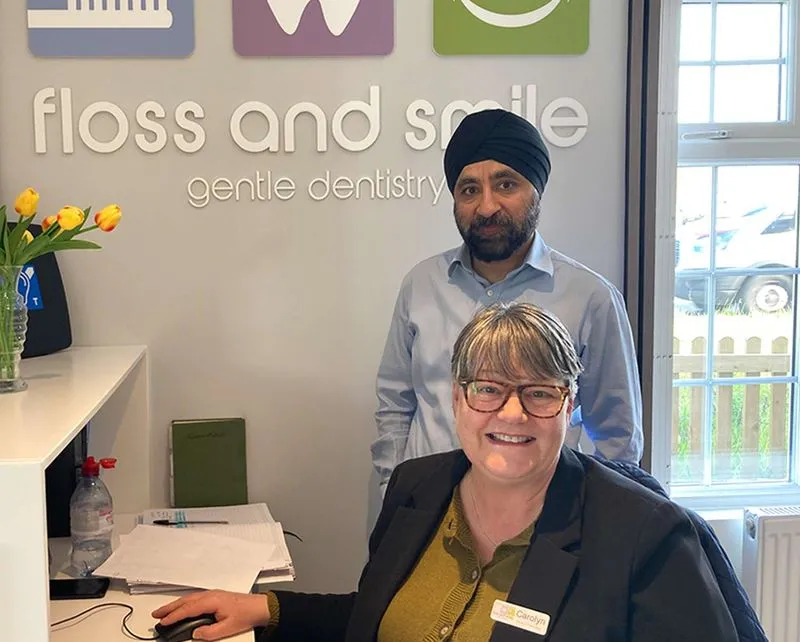 Smiling woman with glasses and a name tag seated at a desk, with a man wearing a turban standing behind her in a dental office.