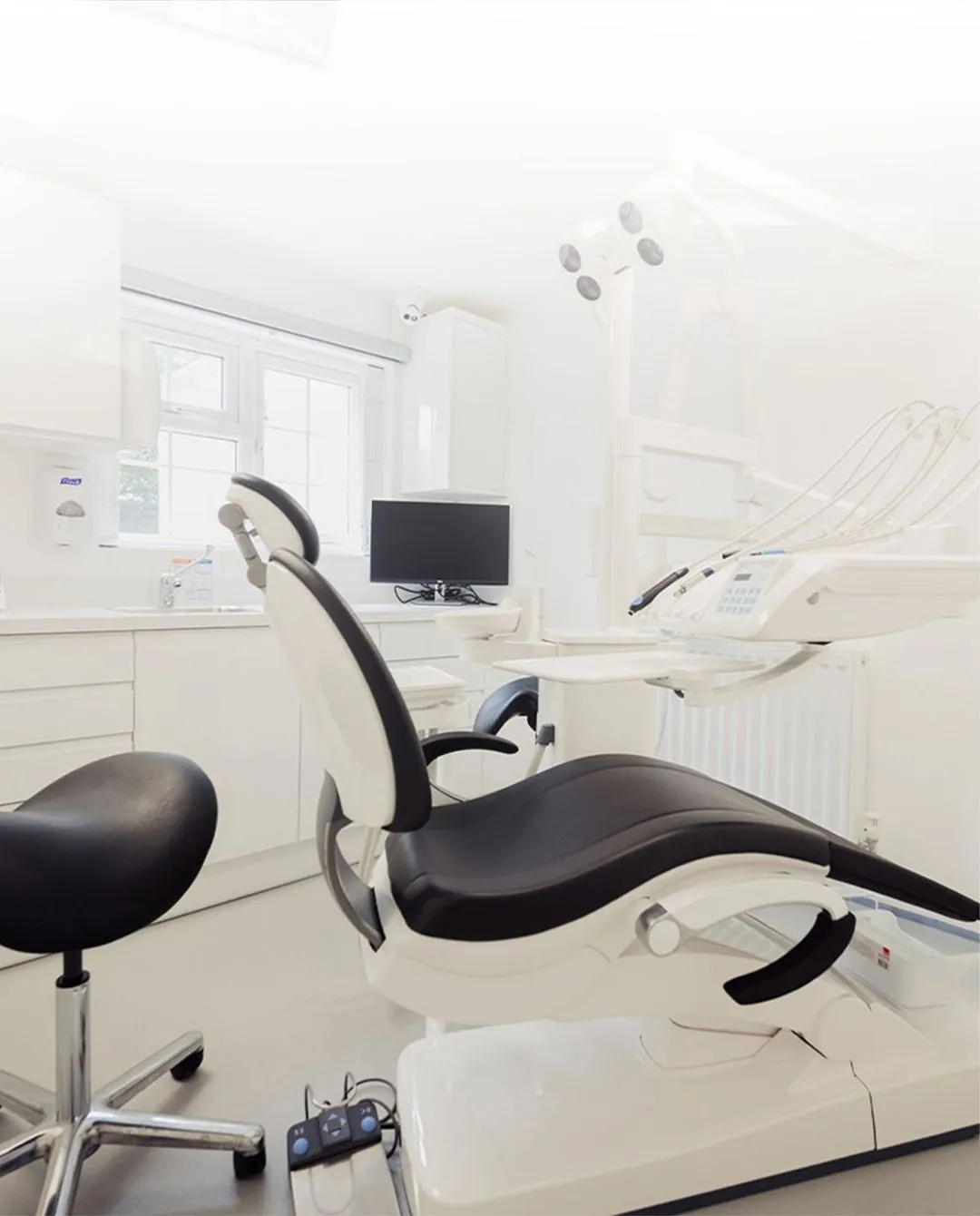 Modern dental clinic room featuring a black and white dental chair, a black stool, and dental equipment against white walls and cabinets.