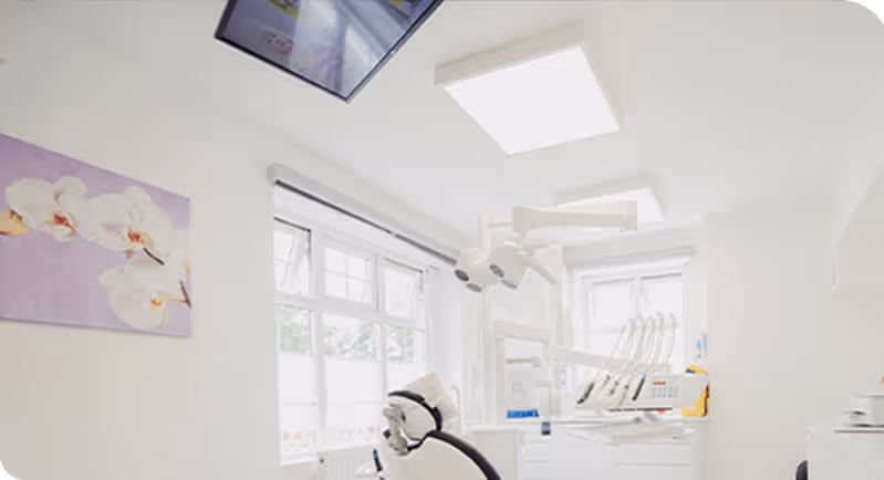 Bright dental clinic room with a dental chair, overhead dental lights, and a floral painting on the wall.