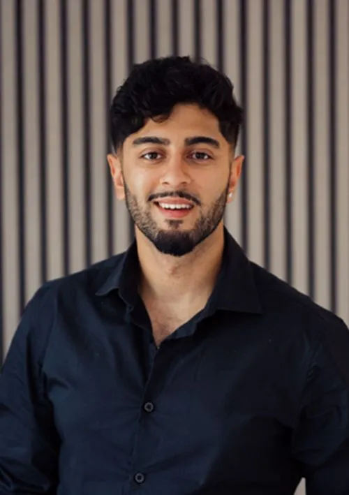 Smiling man with dark hair and beard wearing a black button-up shirt standing in front of a striped background.