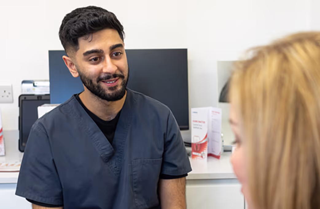 Smiling male healthcare professional in navy scrubs talking to a blonde woman in a medical office.