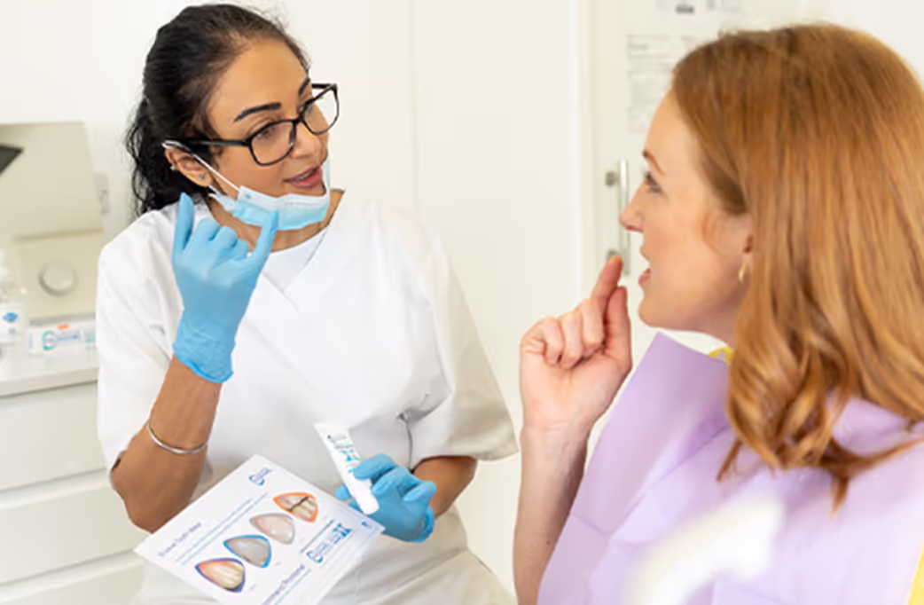 Dentist wearing glasses and gloves explaining dental care with a toothpaste tube and tooth diagram to a patient in a dental office.