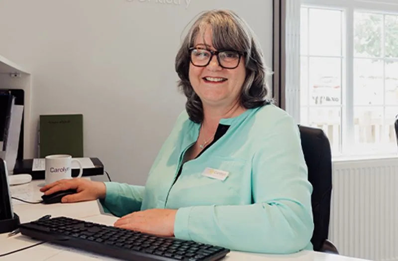 Smiling woman with glasses wearing a light blue blouse sitting at a desk with a keyboard and mouse in an office with a window.