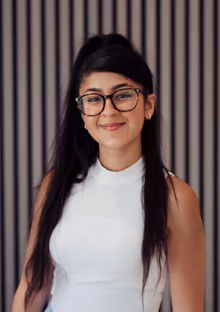 Smiling woman with long dark hair wearing glasses and a white sleeveless top in front of a striped background.