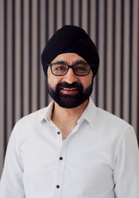 Smiling man wearing glasses, a black turban, and a white button-up shirt standing in front of a striped background.