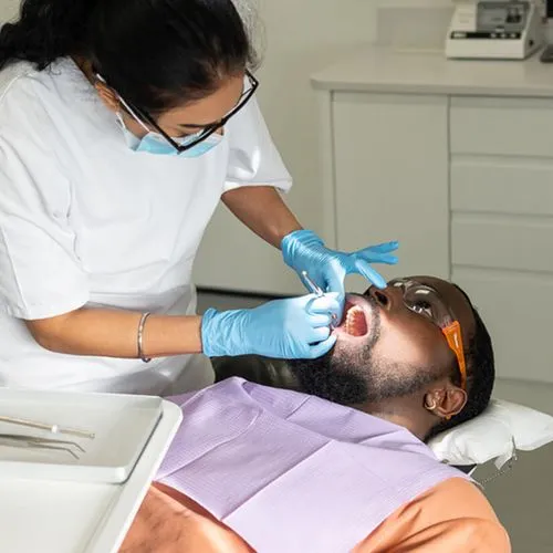 Dentist wearing blue gloves and mask examining a male patient's teeth in a dental clinic.