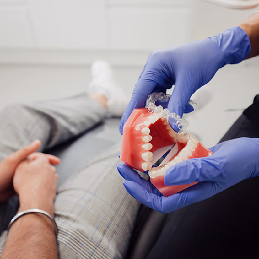 Dentist wearing blue gloves holding a dental model with clear aligners, with a patient seated in the background.