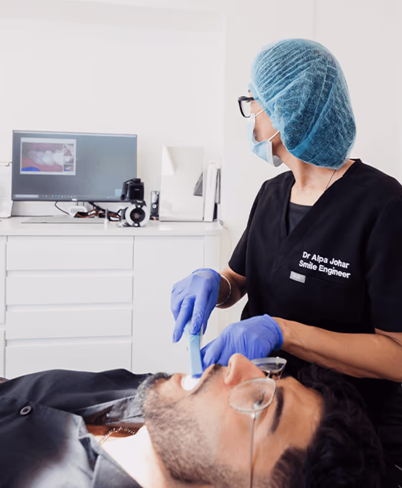 Dentist wearing blue gloves and surgical cap applying dental treatment to a male patient lying back in a dental chair.