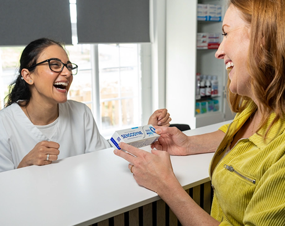 Smiling woman in yellow jacket holding Sensodyne toothpaste while interacting with a laughing female pharmacist behind the counter.