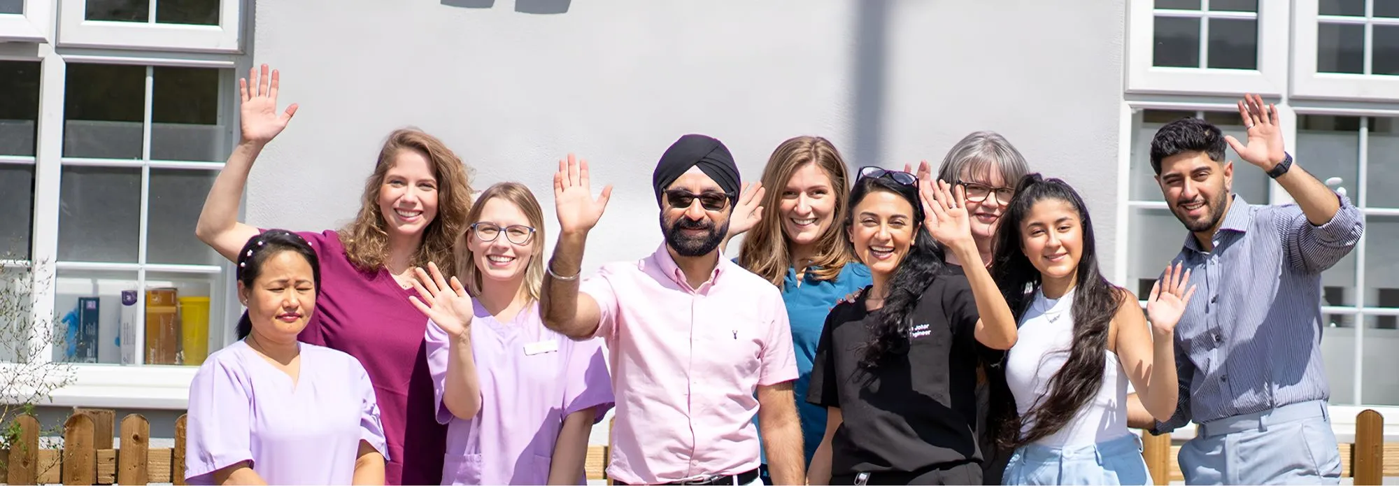 Diverse group of nine smiling people standing outdoors waving at the camera in front of a building with windows.