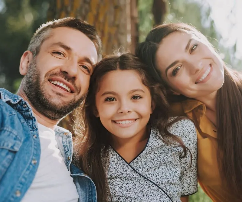 Smiling father, daughter, and mother posing closely together outdoors in natural sunlight.
