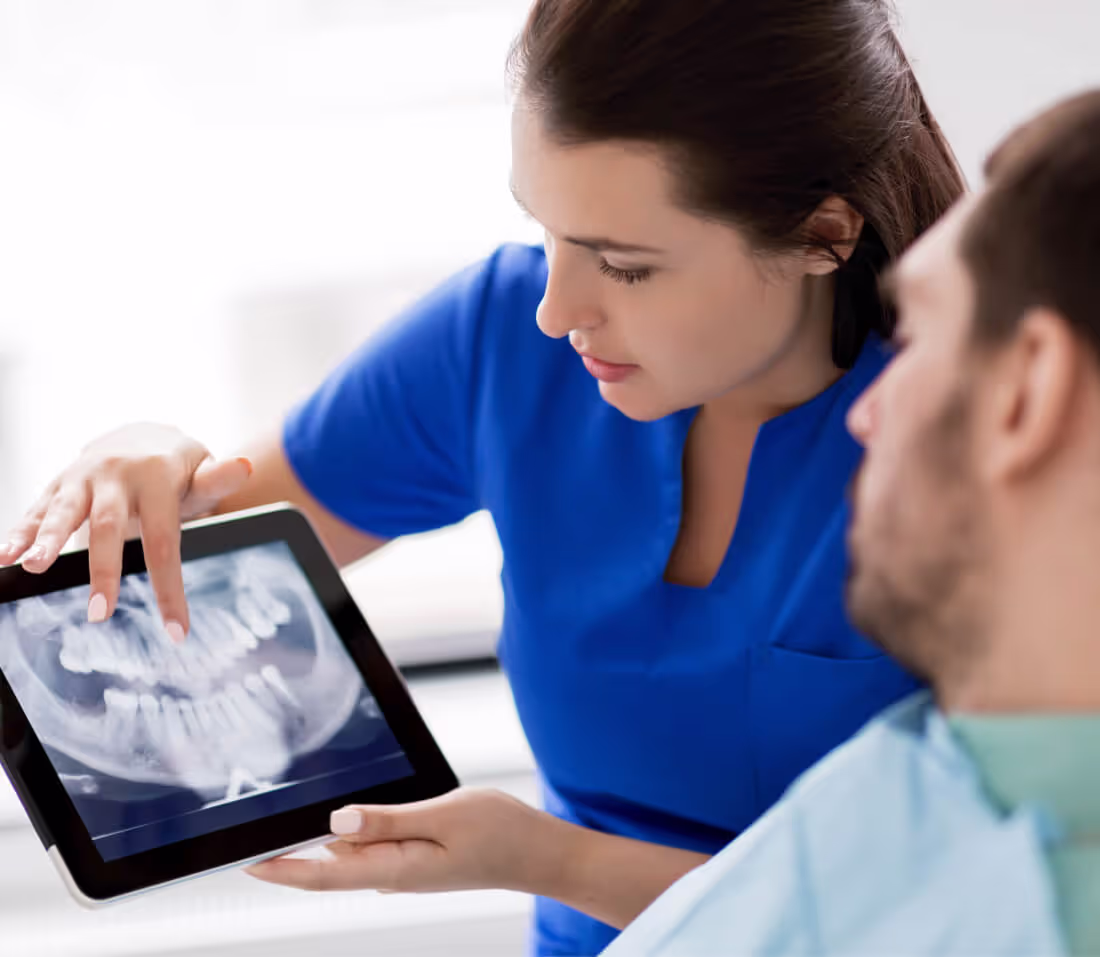 Dental professional in blue scrubs showing a patient's dental X-ray on a tablet.