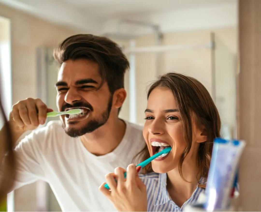 Smiling young man and woman brushing their teeth together in a bathroom mirror.