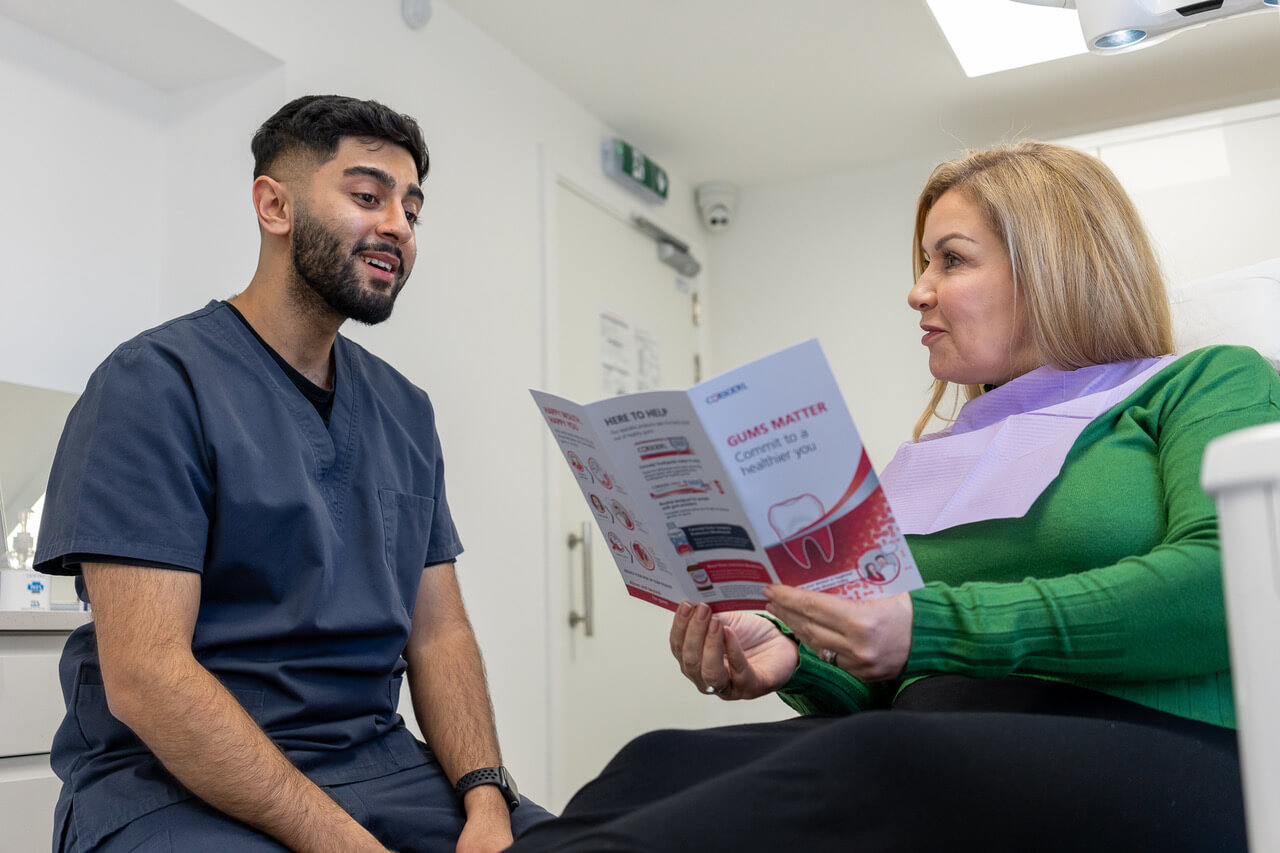 Dentist in navy scrubs talking to a seated woman in a green sweater holding a dental brochure inside a dental office.