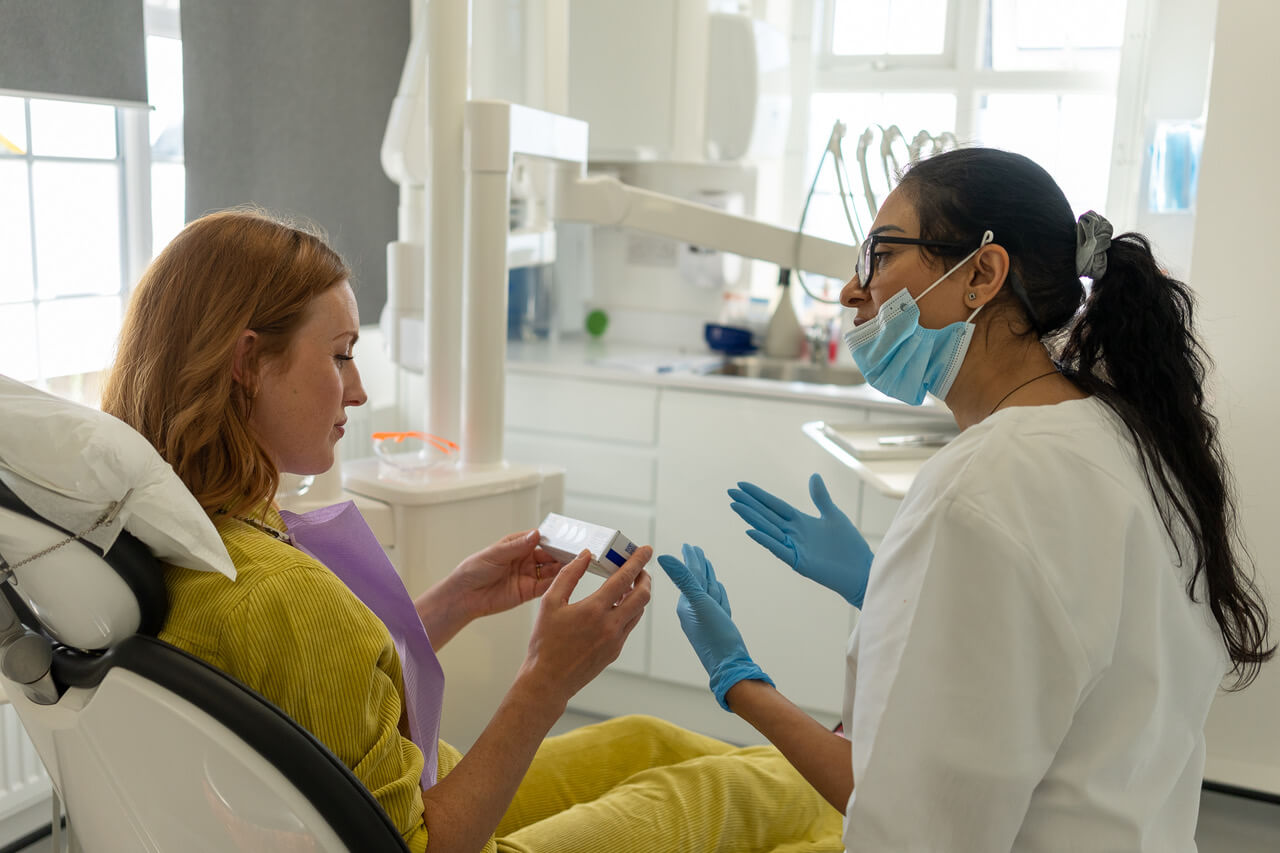 Dentist wearing blue gloves and face mask talking to a female patient holding a dental product in a clinic.