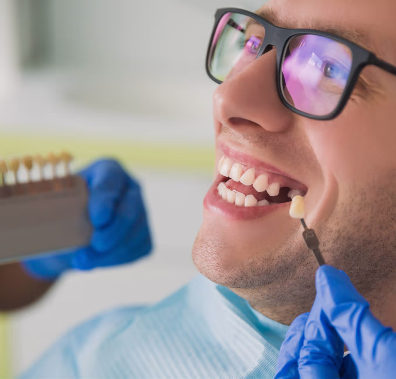Dentist matching a dental veneer shade to a smiling man's teeth using a shade guide.