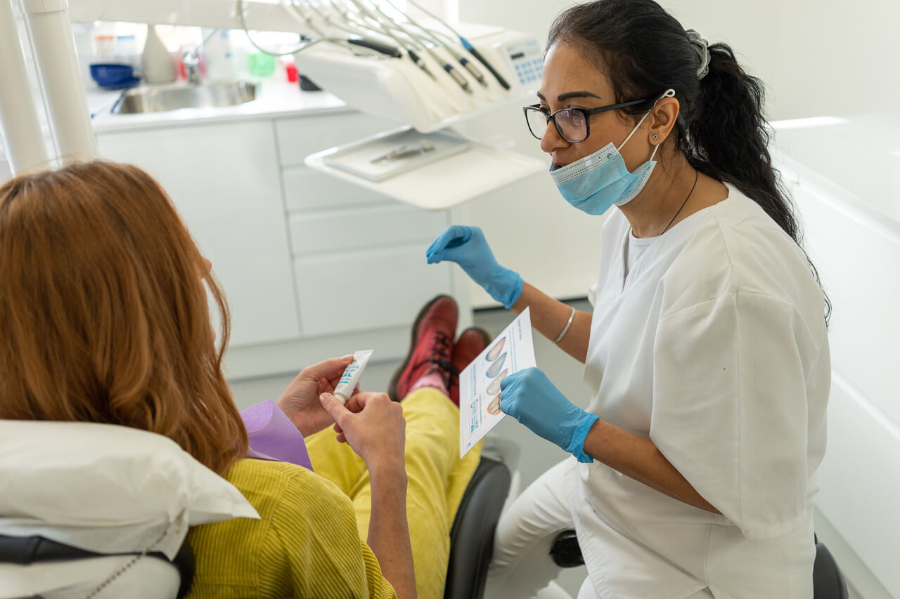Dentist wearing a mask and gloves showing a dental care leaflet to a patient seated in a dental chair.