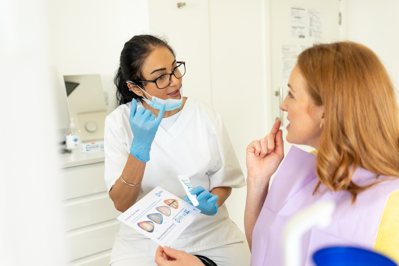 Dentist in white coat and blue gloves explaining dental treatment using a pamphlet to a patient wearing a purple bib.