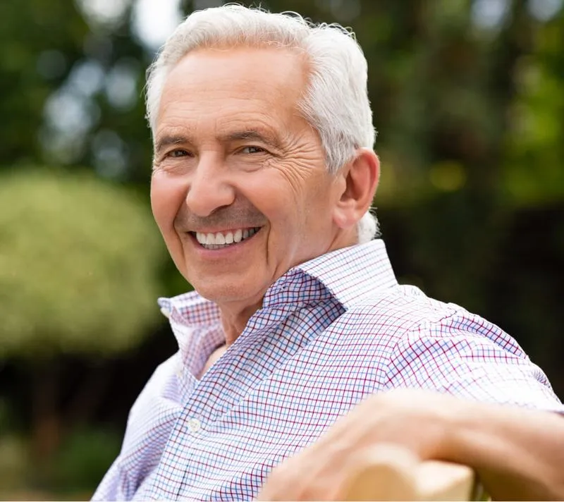 Smiling elderly man with white hair wearing a checkered shirt sitting outdoors.