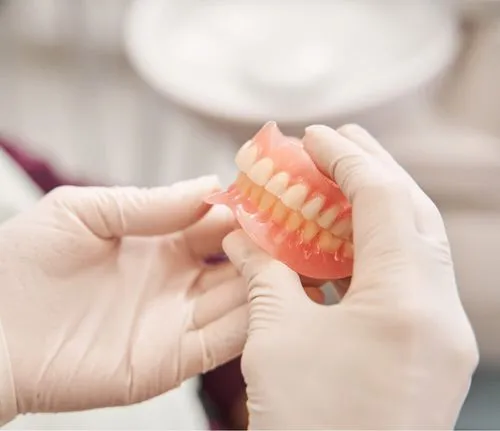 Hands wearing white medical gloves holding a set of pink dentures with white teeth.