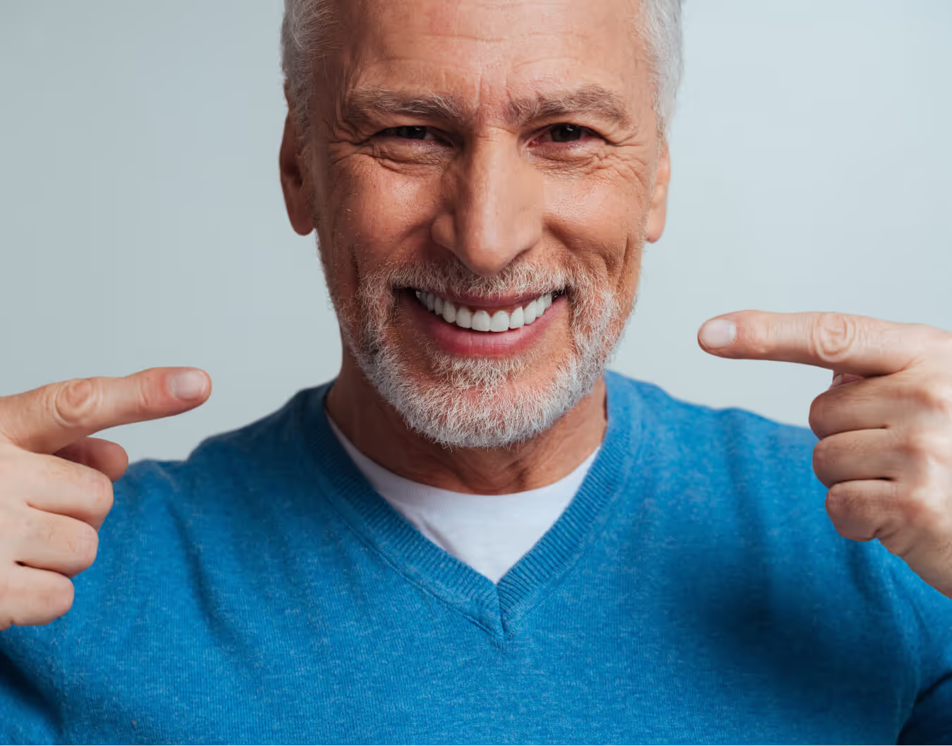 Smiling elderly man with white beard pointing at his white teeth while wearing a blue sweater.