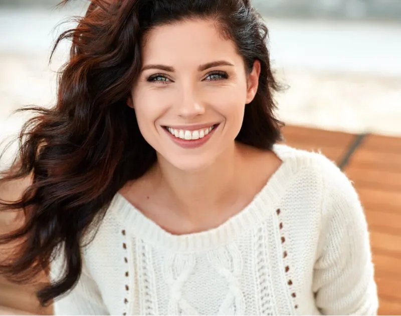 Smiling woman with long wavy brown hair wearing a white knit sweater.