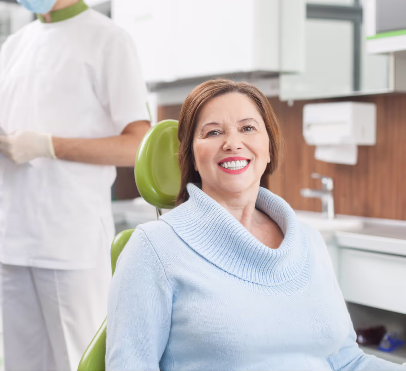 Smiling middle-aged woman sitting in a dental chair with a dentist in the background.