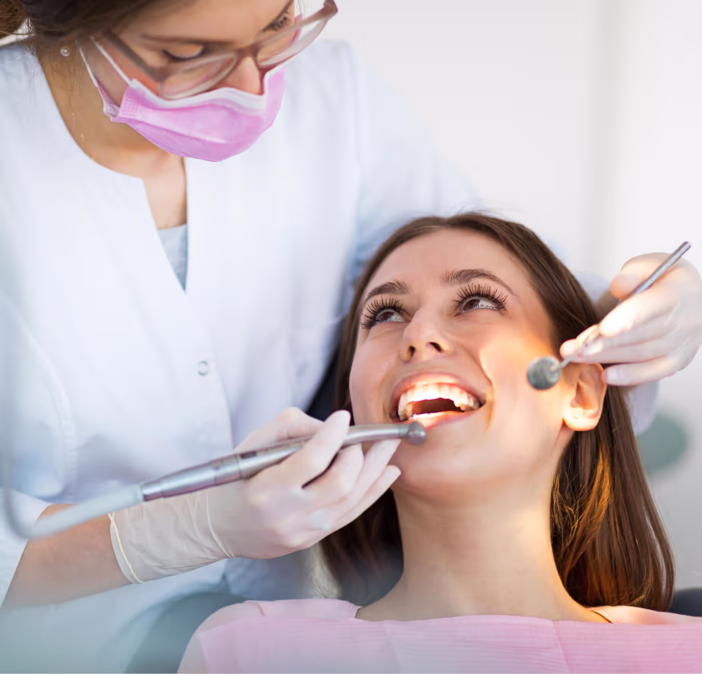 Dentist wearing glasses and pink mask examining a smiling woman’s teeth with dental tools.