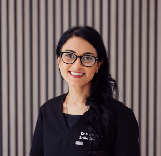 Smiling woman with long dark hair and glasses wearing a black medical uniform standing in front of a vertical striped wall.