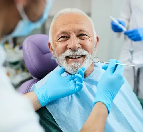 Smiling elderly man with white beard receiving dental checkup from dentist wearing blue gloves and mask.