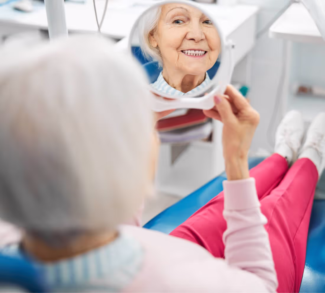 Senior woman sitting in a dental chair smiling while looking at her teeth in a handheld mirror.