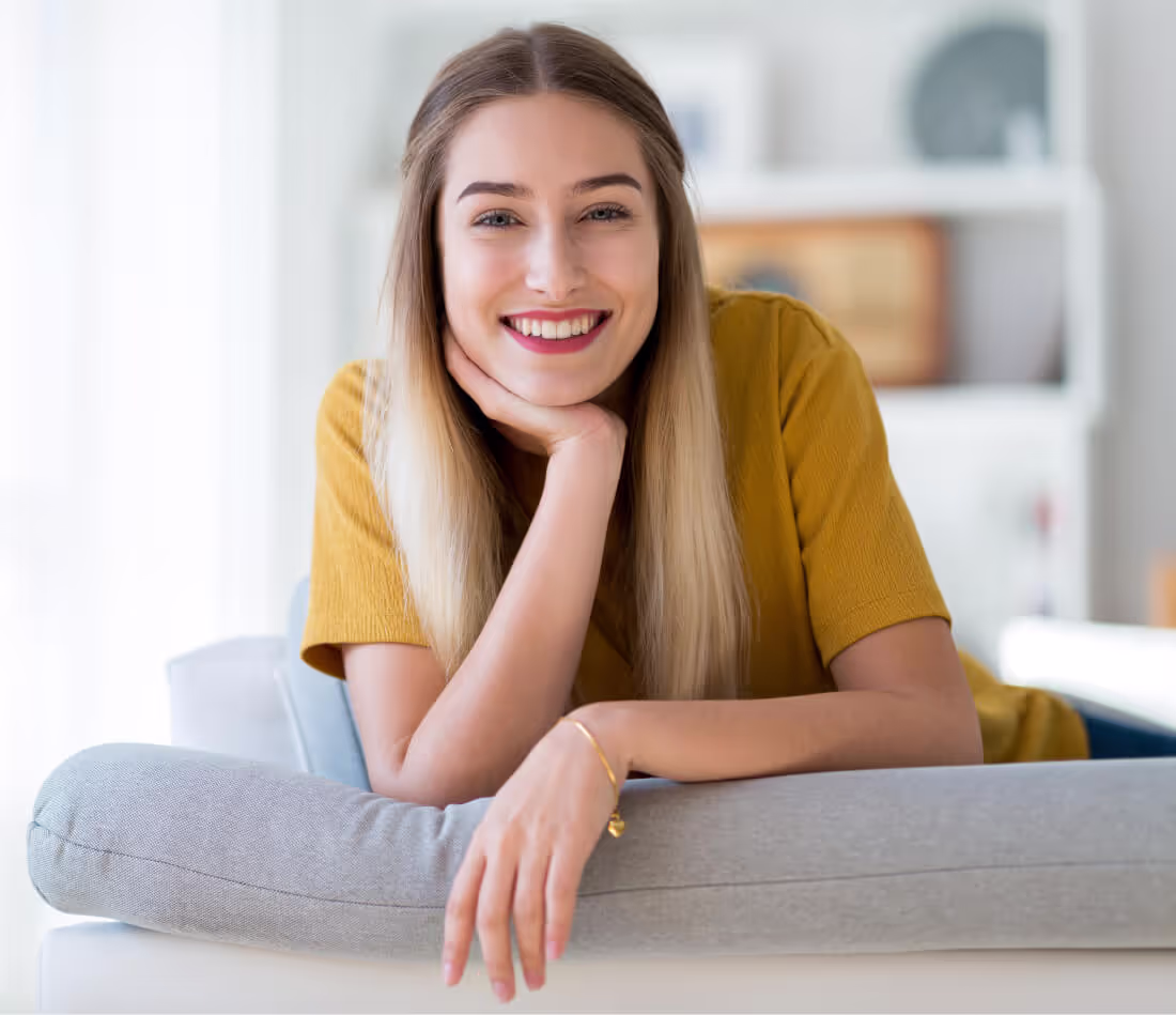 Smiling woman with long blond hair wearing a mustard yellow top, leaning on a gray sofa cushion in a bright room.