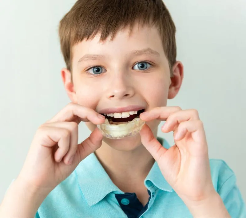 Boy with blue eyes and brown hair holding a clear dental aligner near his mouth.