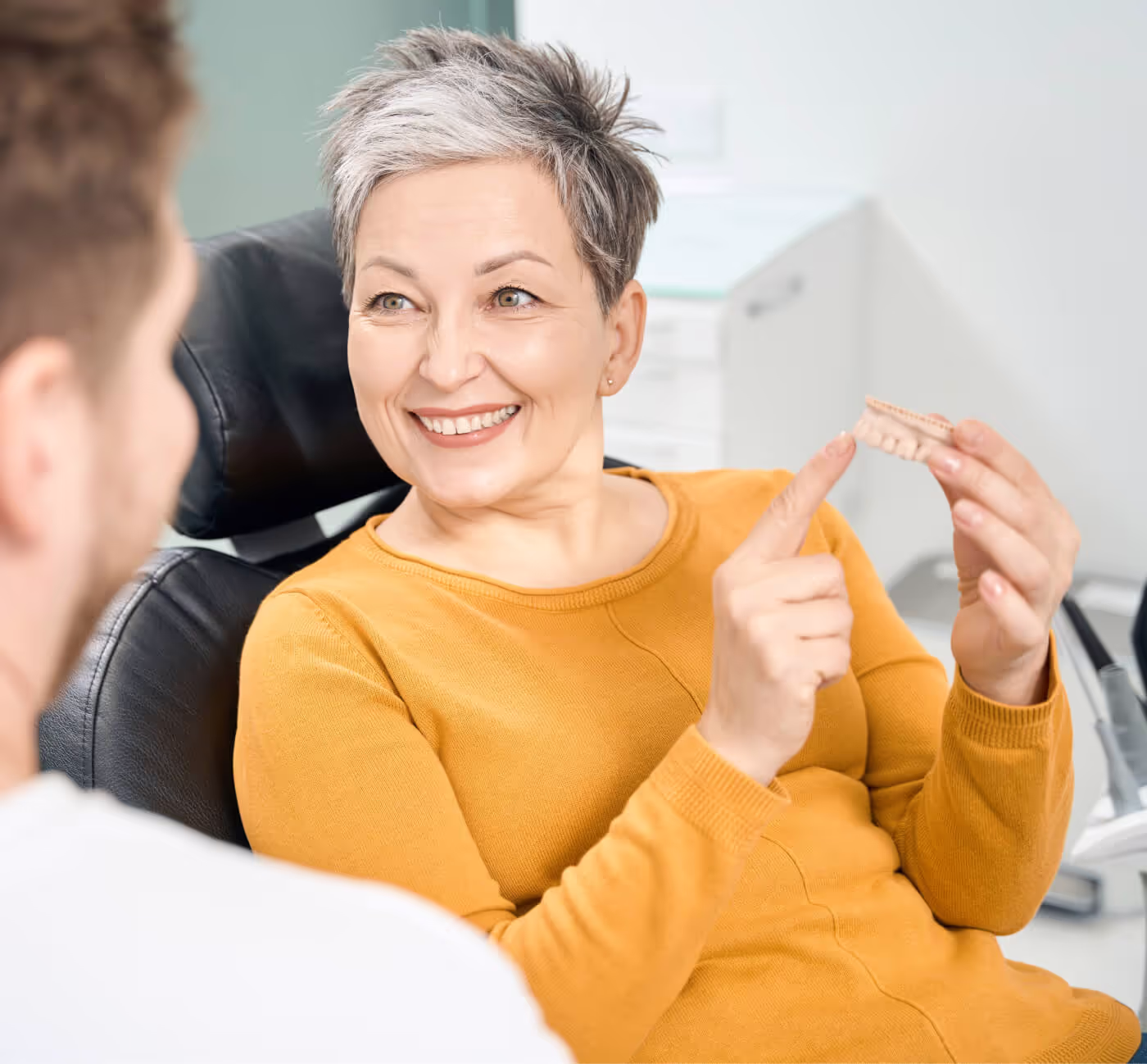 Smiling woman with short gray hair sitting in a dental chair, holding and pointing to a dental model while talking to a dentist.