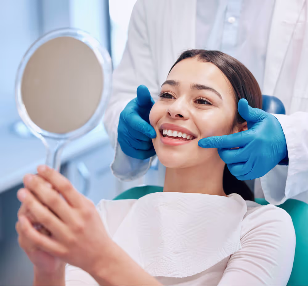 Smiling woman in dental chair holding a mirror while dentist with blue gloves points at her teeth.