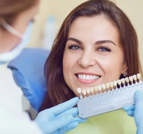 Dentist with blue gloves holding a tooth shade guide next to a smiling woman showing her white teeth.