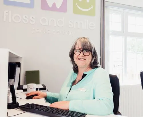 Smiling woman in glasses and turquoise uniform sitting at a reception desk with computer keyboard and mouse.