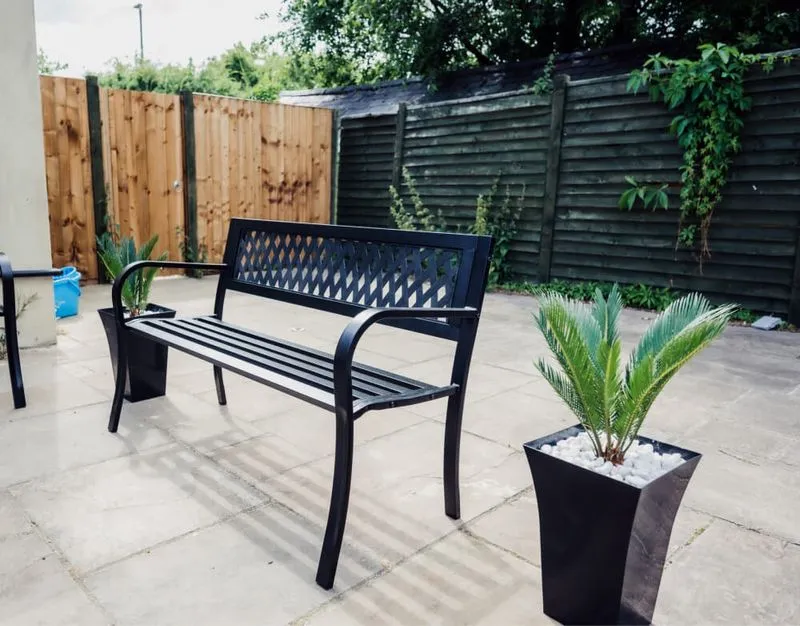 Black metal bench on a stone patio between two tall black planters with green plants, wooden fence background.