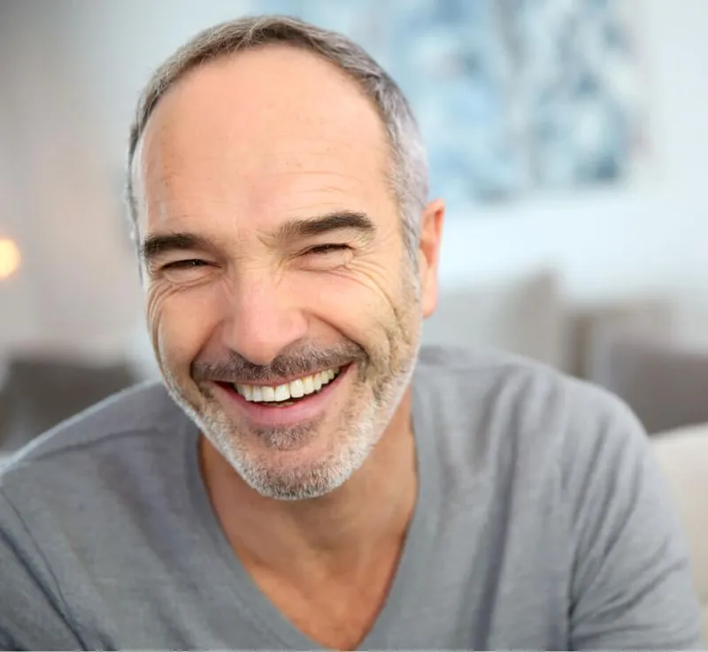 Smiling middle-aged man with gray hair and beard wearing a gray shirt in a bright room.